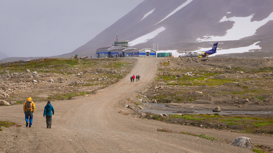 Trekking companions hiking along the road with the Kulusuk airport terminal and Iceland Air Connect plane in the background