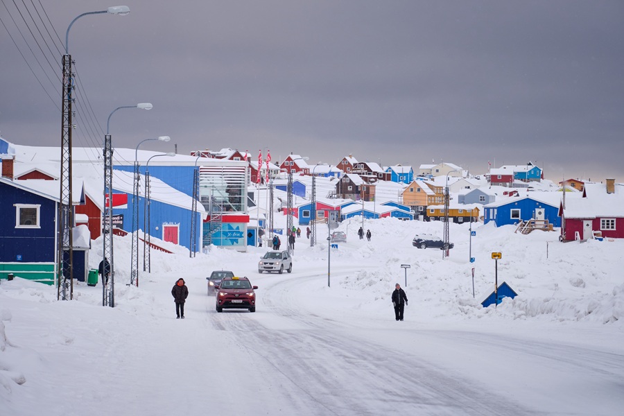 Snow-covered street in a Greenland town with colourful buildings, vehicles, and people moving through winter conditions.