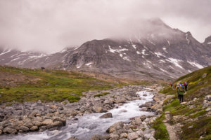 Trekking companions hiking beside a fast-flowing stream with fog-shrouded mountains in the background