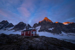 Sunset bathing the tips of the peaks behind the Tasiilaq Mountain Hut with a golden light. The hut is in the foreground