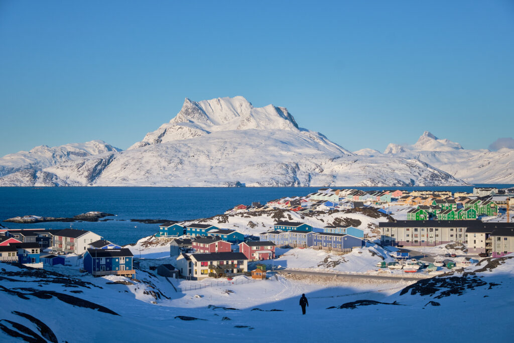 Coastal Greenland settlement in winter with colourful houses, snow-covered terrain, and mountains across the sea.